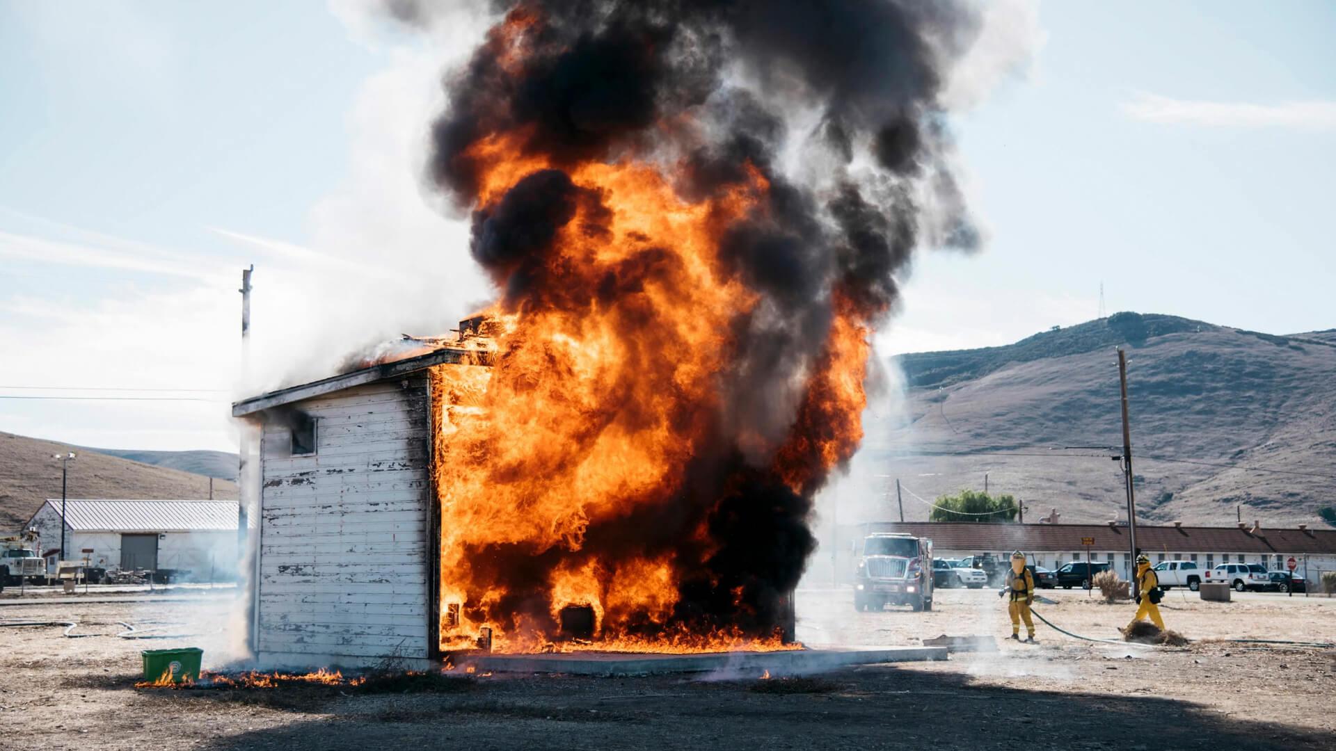 Small building engulfed in flames during a controlled burn test as firefighters observe nearby.