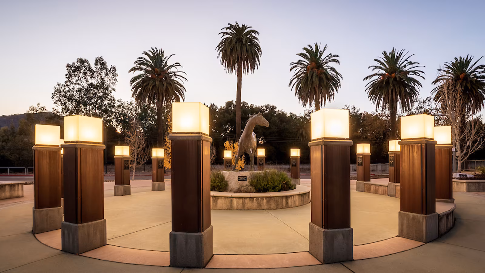 Campus plaza at dusk featuring illuminated columns and a horse statue surrounded by palm trees.